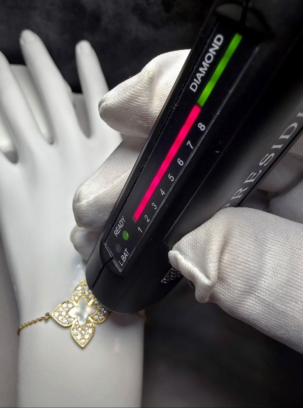 A close-up of a gold butterfly bracelet with a pearl center and small clear stones, shown on a white mannequin wrist while a black gemstone tester device is held over it. White-gloved hands are visible, and the background is dark.