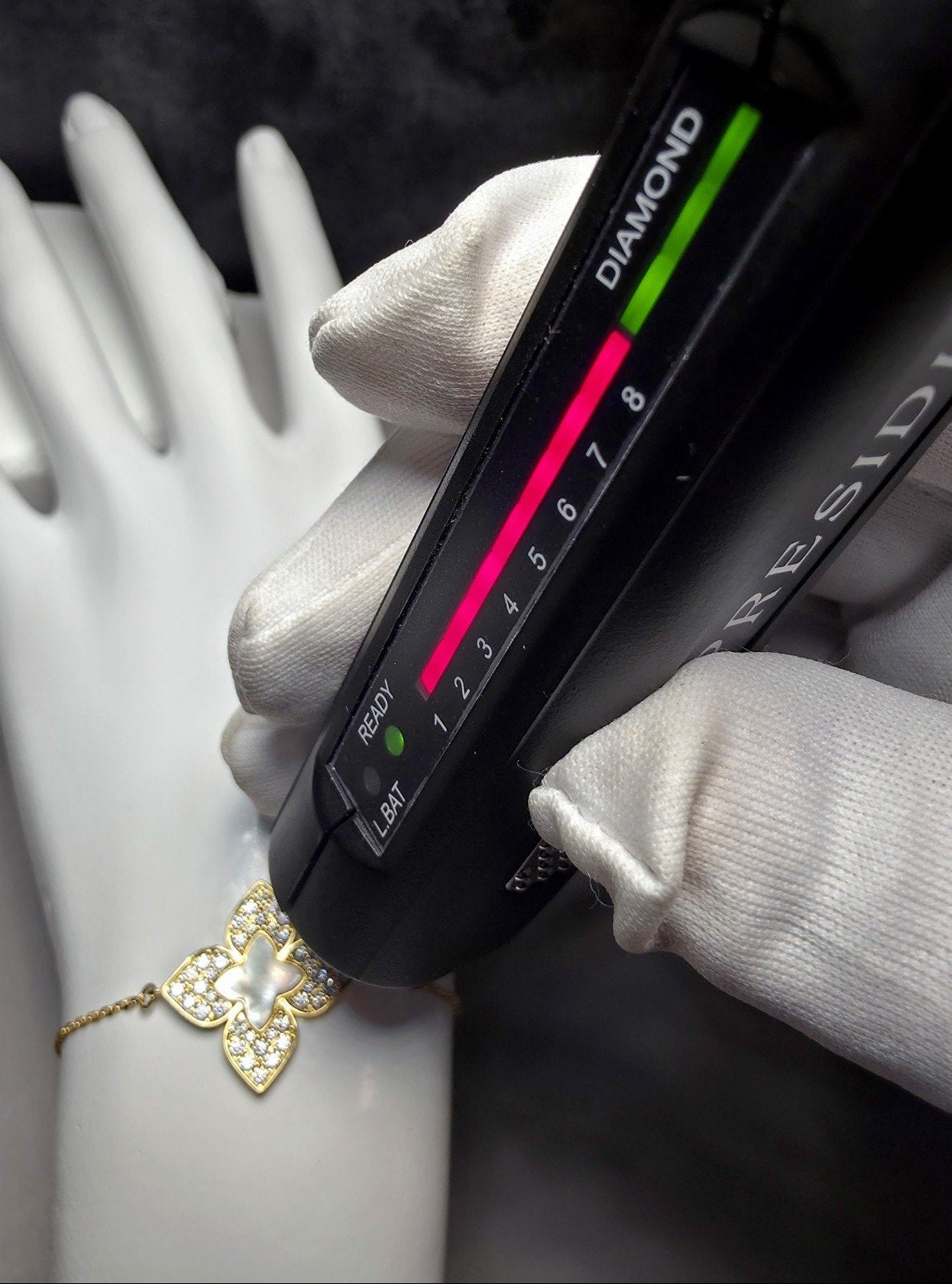 A close-up of a gold butterfly bracelet with a pearl center and small clear stones, shown on a white mannequin wrist while a black gemstone tester device is held over it. White-gloved hands are visible, and the background is dark.