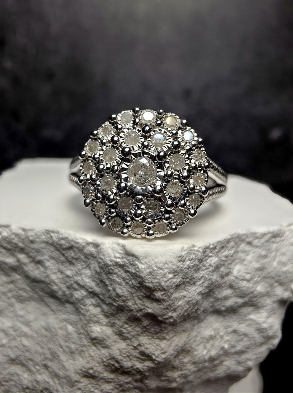 A silver-toned vintage-style cluster ring with multiple round-cut stones, displayed on a textured white stone pedestal against a dark gray background.