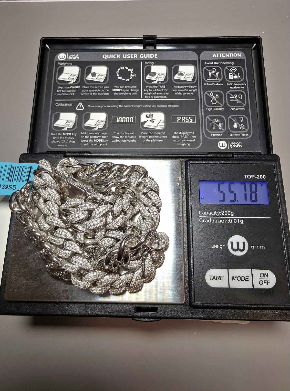 A silver Cuban link chain covered in small clear stones, displayed on a digital scale with its lid open. The scale shows a weight of 55.