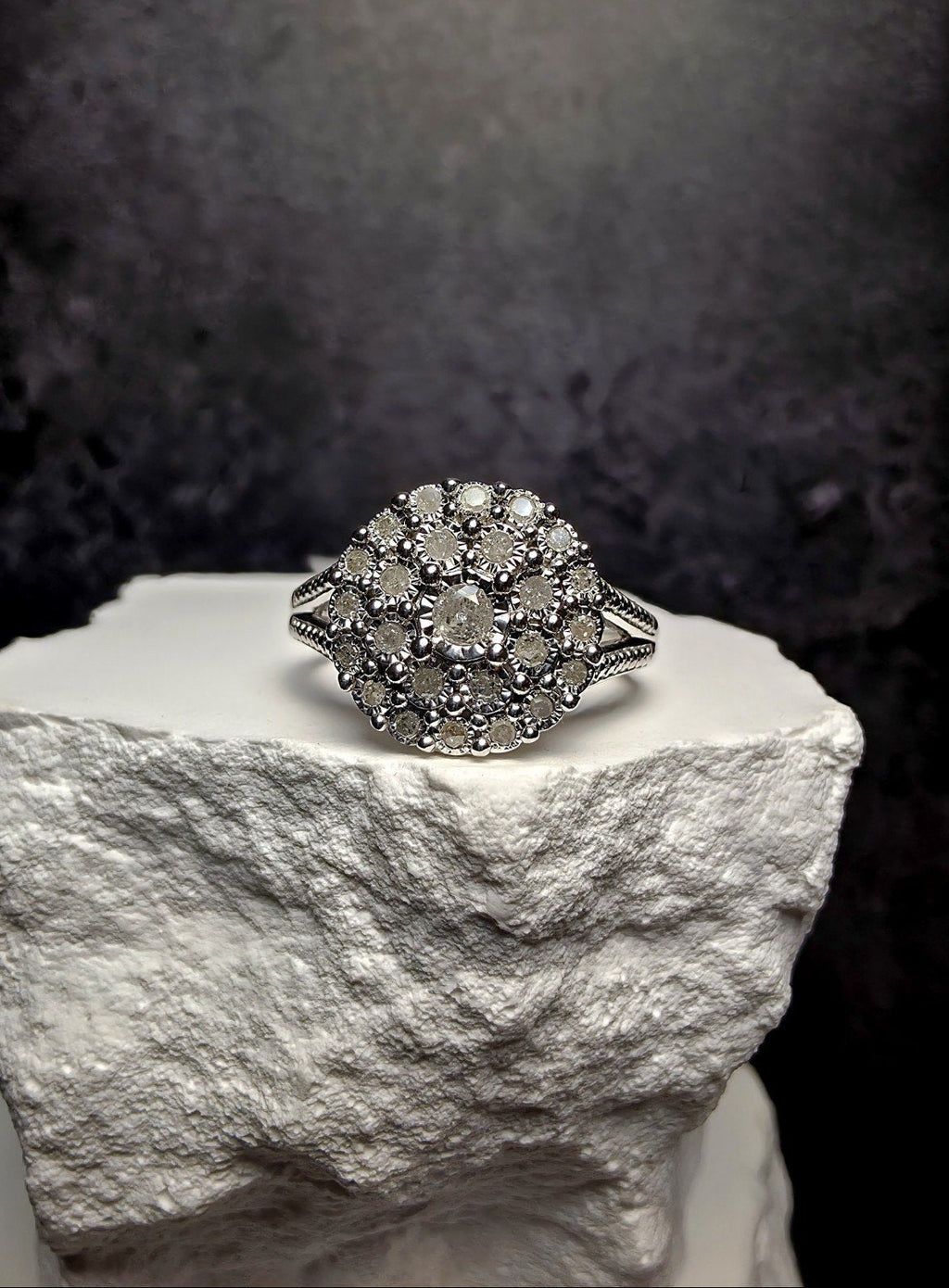 A silver-toned ring with a large round cluster of clear gemstones, displayed on a textured white stone pedestal against a dark gray background.