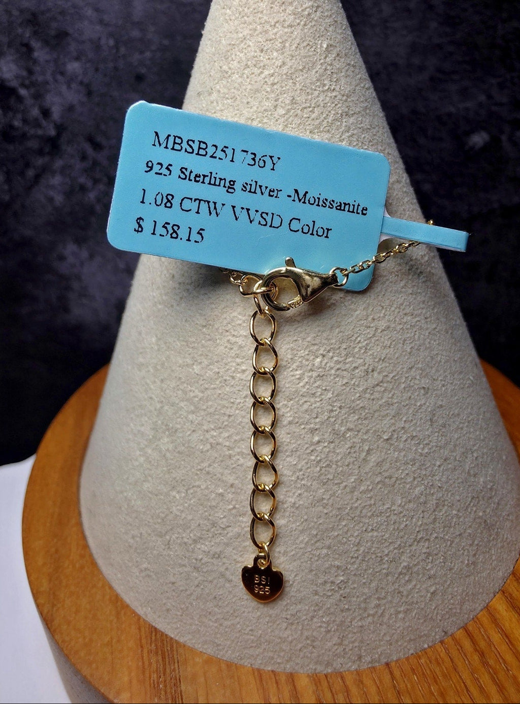 A sterling silver bracelet with a gold-tone extender chain and a small heart-shaped Moissanite charm, displayed on a textured cone stand with a blue product tag.