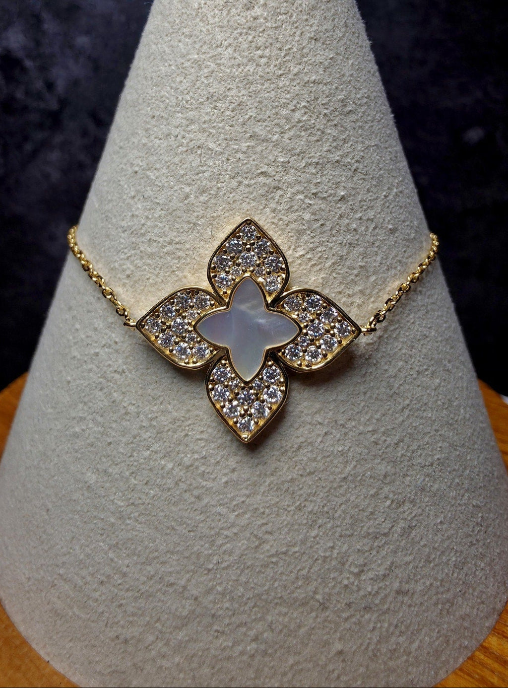 A gold-tone floral bracelet with a mother-of-pearl center and pavé-set clear stones, displayed on a textured light gray jewelry cone.