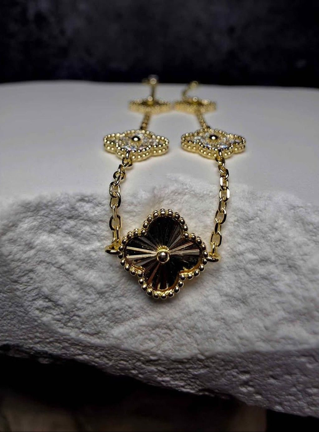 A gold-tone floral chain necklace with a large four-petal flower pendant featuring a black center and clear rhinestone accents, displayed on a textured white stone surface.