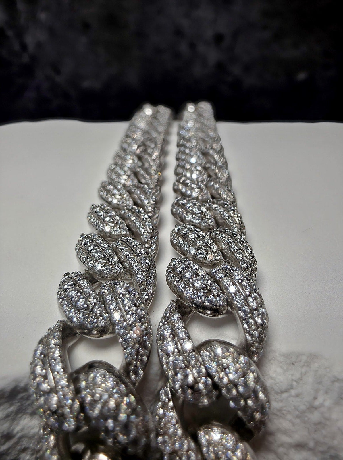 A close-up of a silver-toned Cuban link chain bracelet heavily embellished with clear pavé stones, resting on a white surface with a dark textured background.