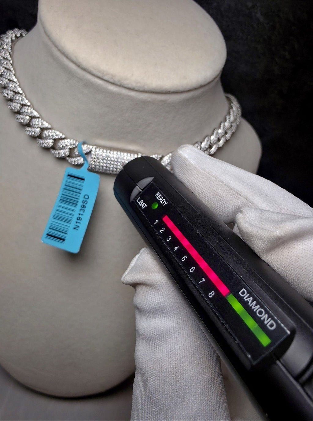 A close-up of a silver-toned diamond necklace displayed on a beige jewelry bust, with a gloved hand holding a black fluorescence test light over the necklace. A blue inventory tag is attached to the necklace.