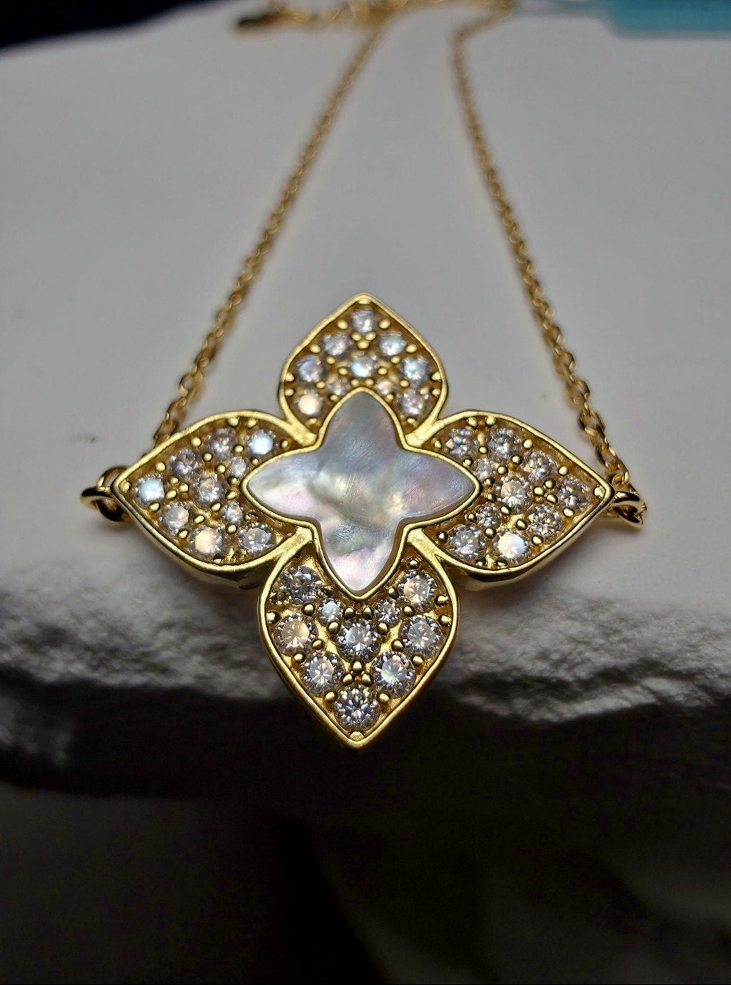 A gold-tone floral pendant necklace featuring a central iridescent mother-of-pearl star surrounded by clear pavé stones, displayed on a textured white surface.