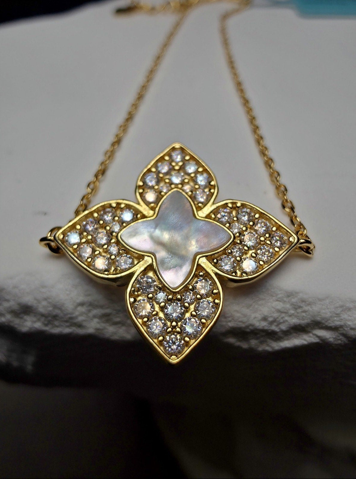 A gold-tone floral pendant necklace featuring a central iridescent mother-of-pearl star surrounded by clear pavé stones, displayed on a textured white surface.