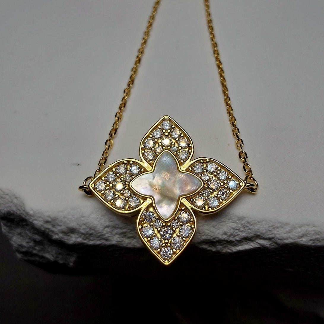 A gold-tone floral pendant necklace featuring a central iridescent mother-of-pearl star surrounded by clear pavé stones, displayed on a dark textured surface against a light gray background.