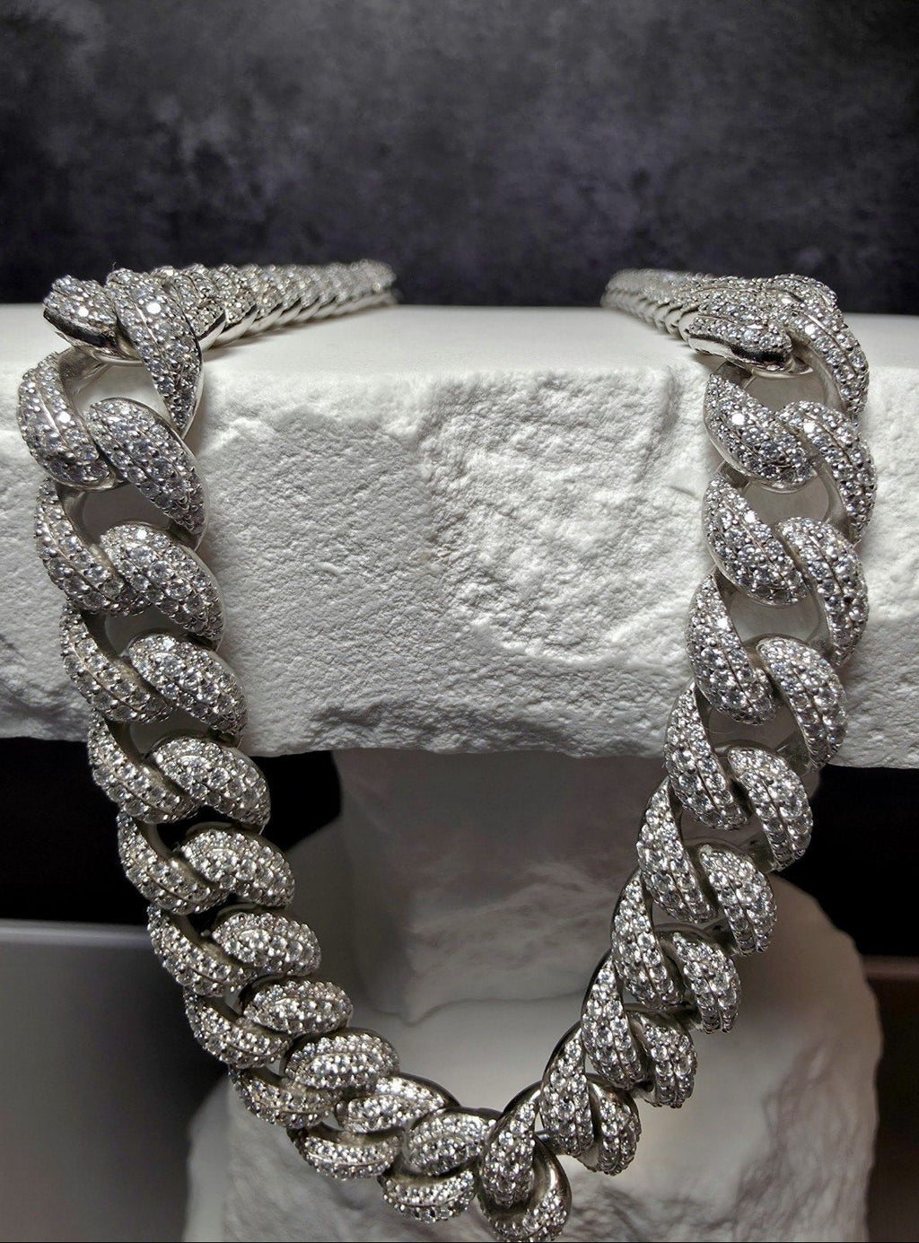A thick silver-toned Cuban link chain necklace, heavily embellished with clear pavé stones, draped over a white textured stone block against a dark gray background.