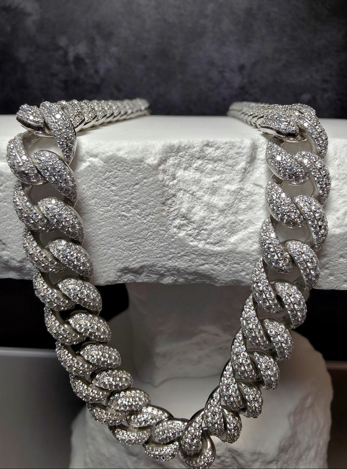 A thick silver-toned Cuban link chain necklace, heavily embellished with clear pavé stones, draped over a white textured stone block against a dark gray background.