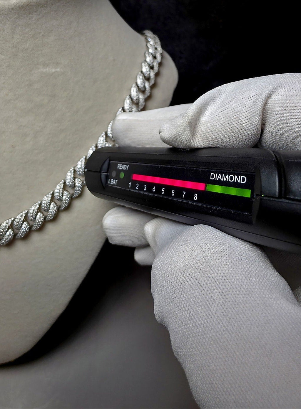 A close-up of a silver diamond-encrusted chain necklace displayed on a beige mannequin bust, with a gloved hand holding a black diamond tester tool against the chain.