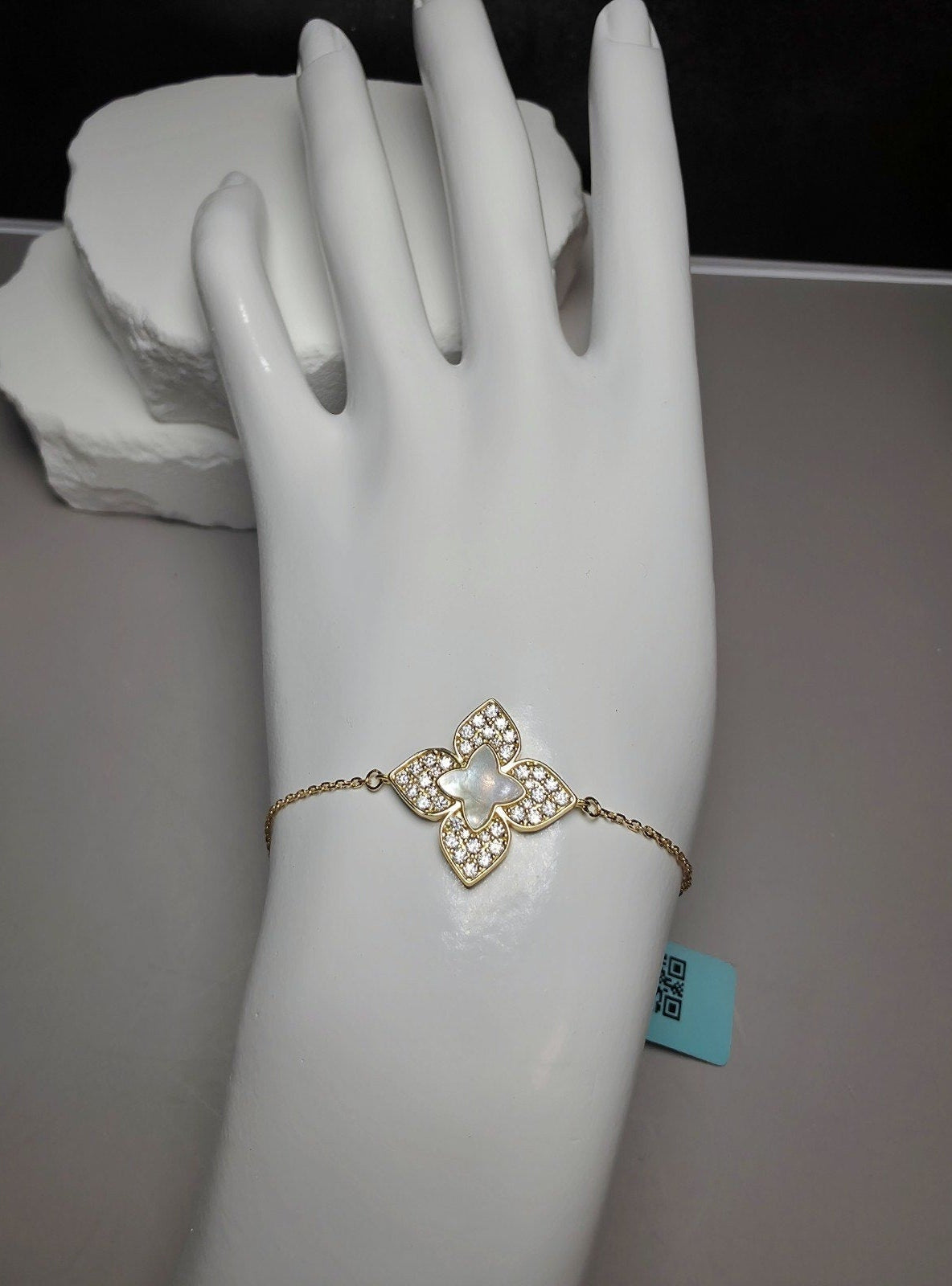 A gold-tone bracelet with a central floral motif set with clear stones and a pearl-like center, displayed on a white mannequin hand. A small turquoise tag is attached near the wrist.