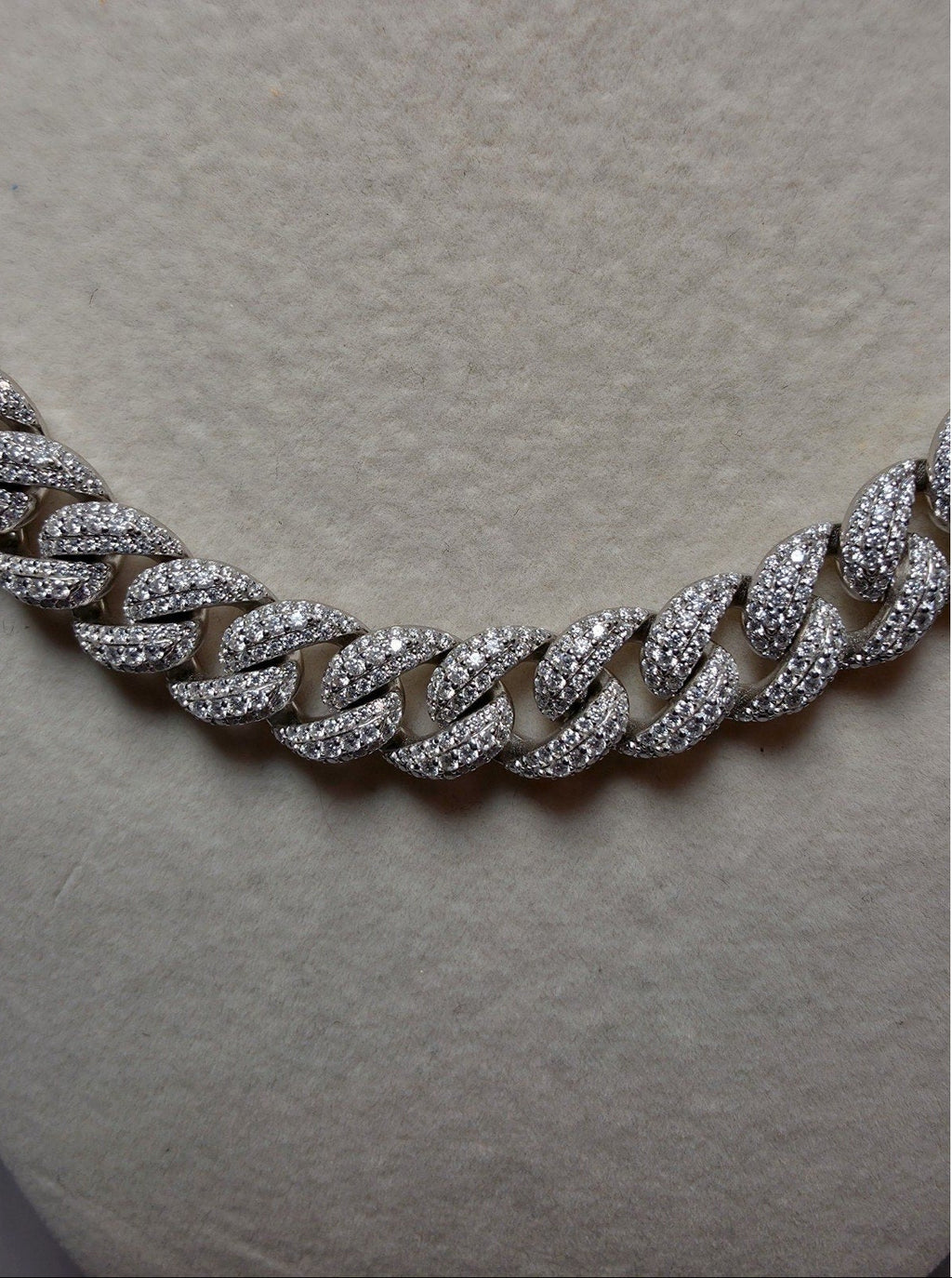 A close-up of a silver-toned Cuban link chain necklace paved with small clear stones, displayed on a textured beige jewelry bust.