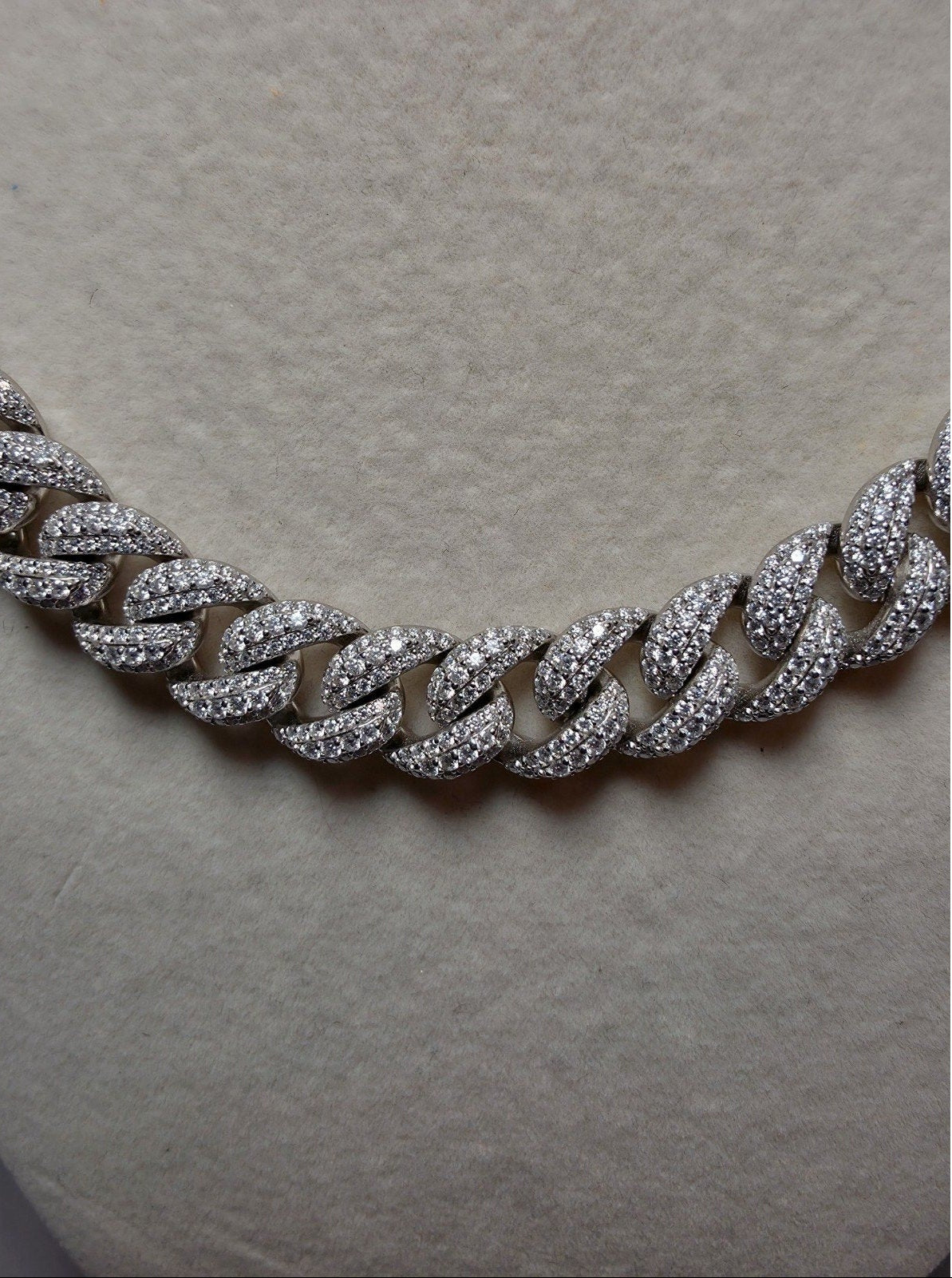 A close-up of a silver-toned Cuban link chain necklace paved with small clear stones, displayed on a textured beige jewelry bust.