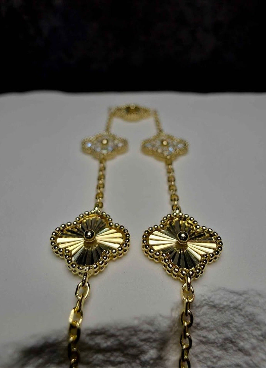 A pair of gold-toned statement earrings with intricate filigree clover motifs and dangling chain links, displayed on a white textured surface against a dark background.