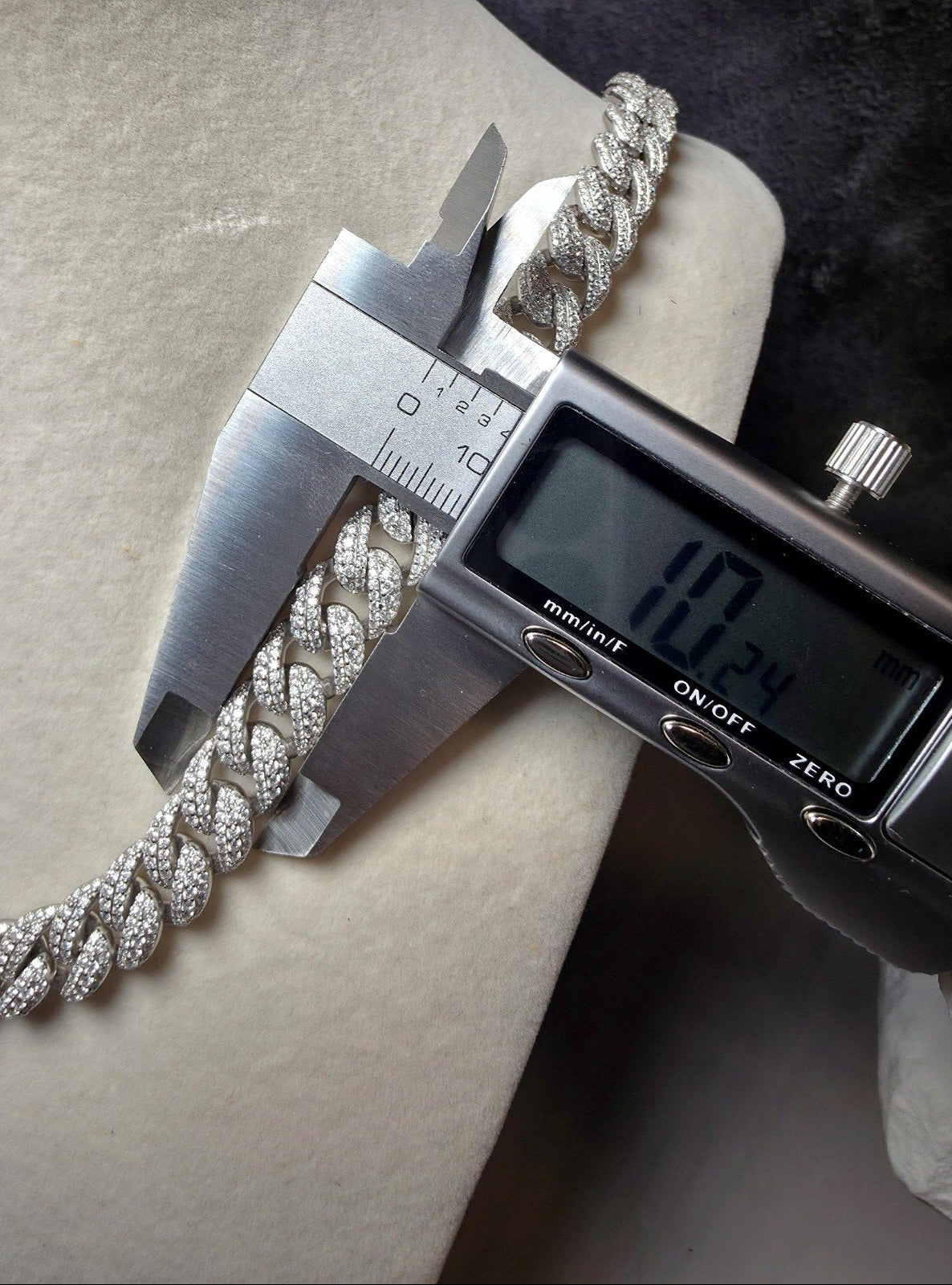 A silver-toned chain bracelet covered in small clear stones, being measured with a digital caliper on a light beige surface.