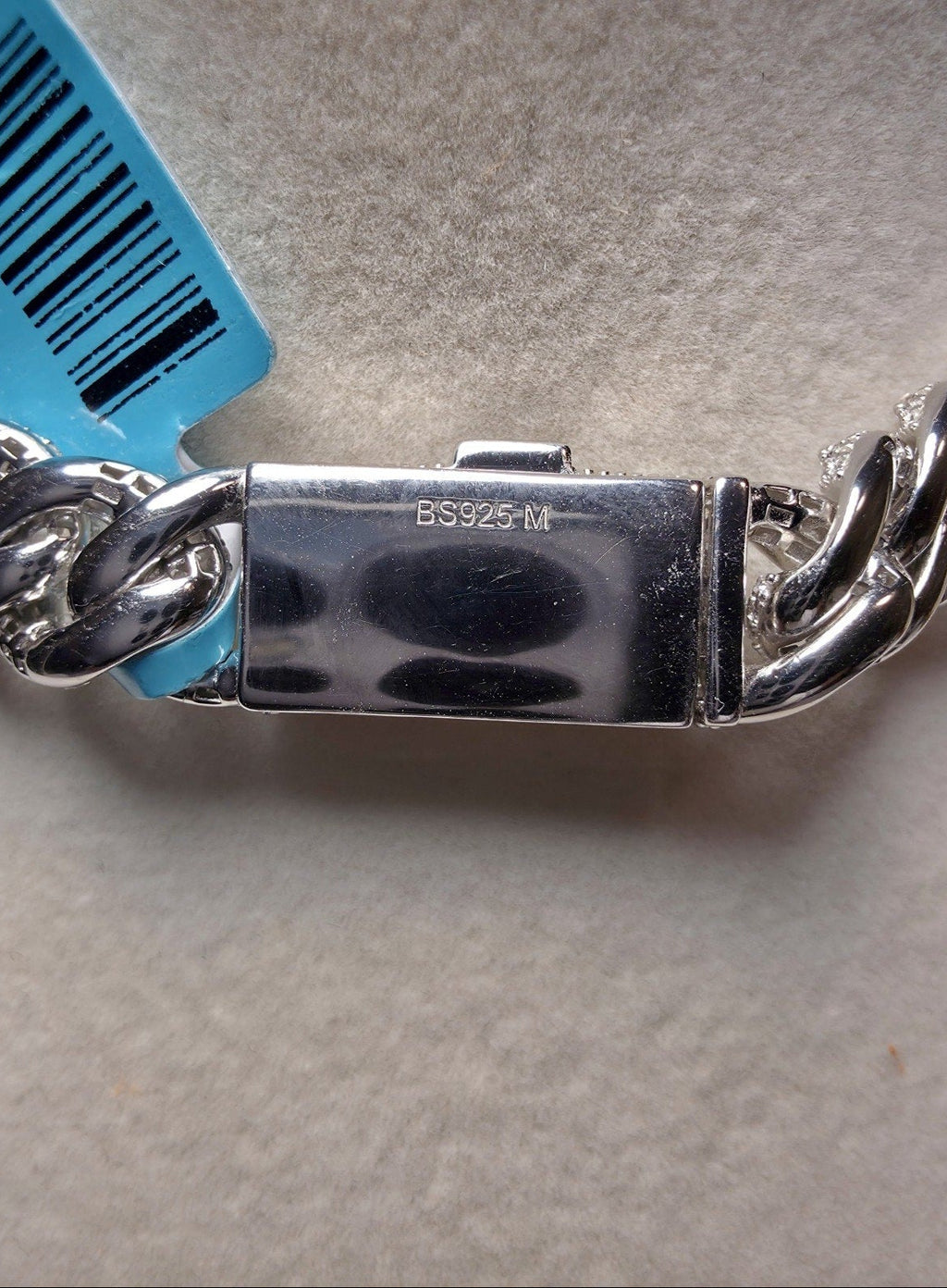 Close-up of a silver-toned metal chain bracelet clasp with a blue barcode tag attached, photographed on a light neutral surface.