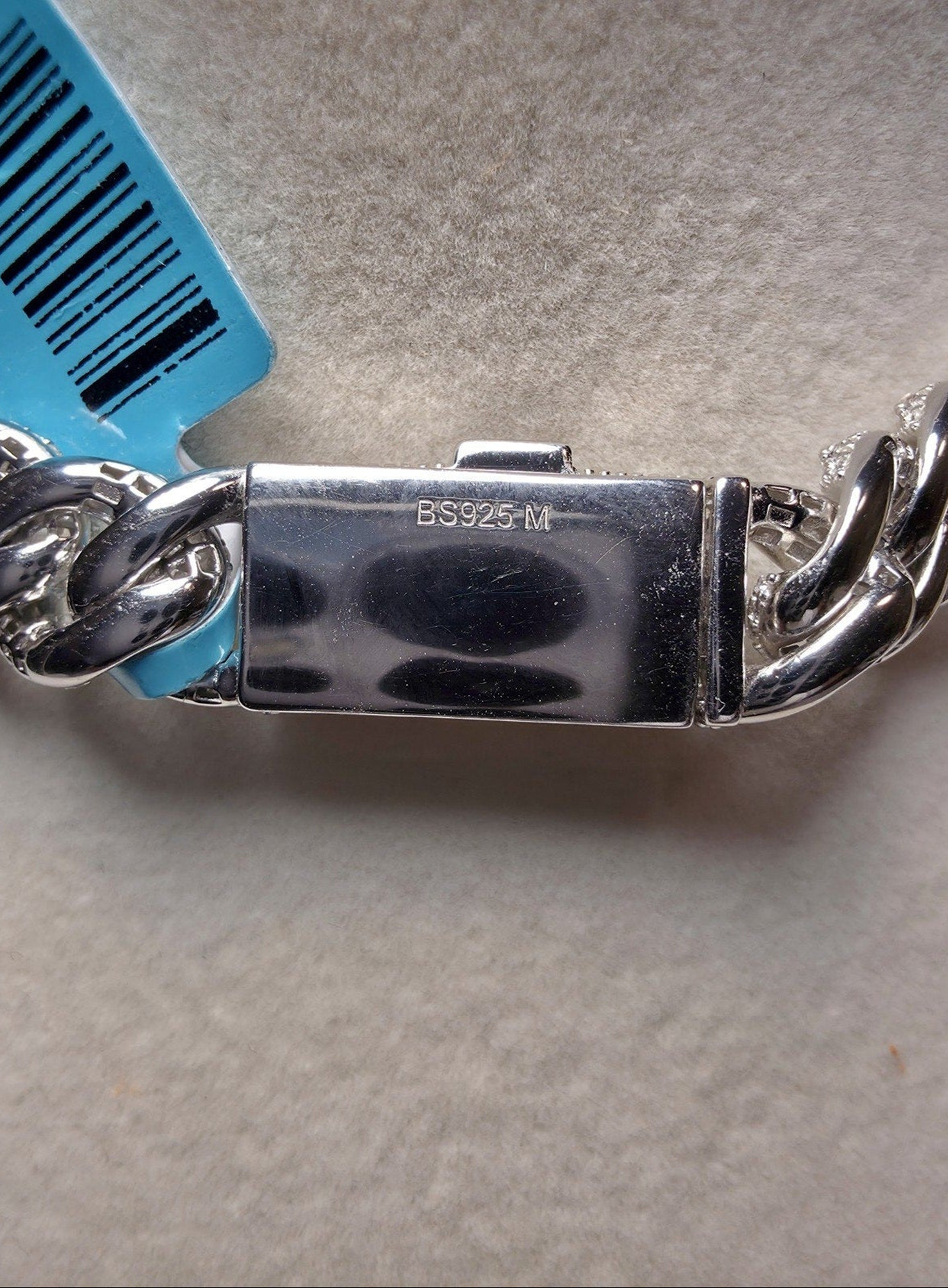 Close-up of a silver-toned metal chain bracelet clasp with a blue barcode tag attached, photographed on a light neutral surface.