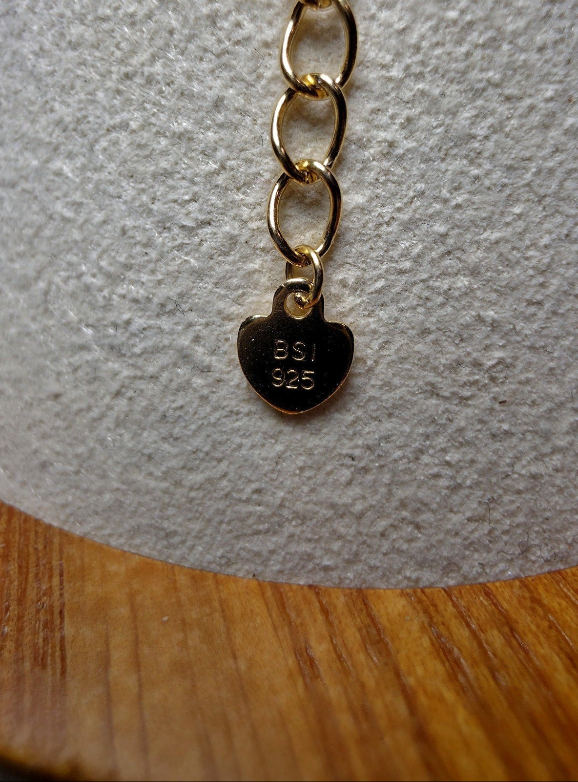 A gold heart-shaped pendant charm engraved with 'BSI 925' on a matching gold chain, displayed on a textured white jewelry display bust over a wooden surface.