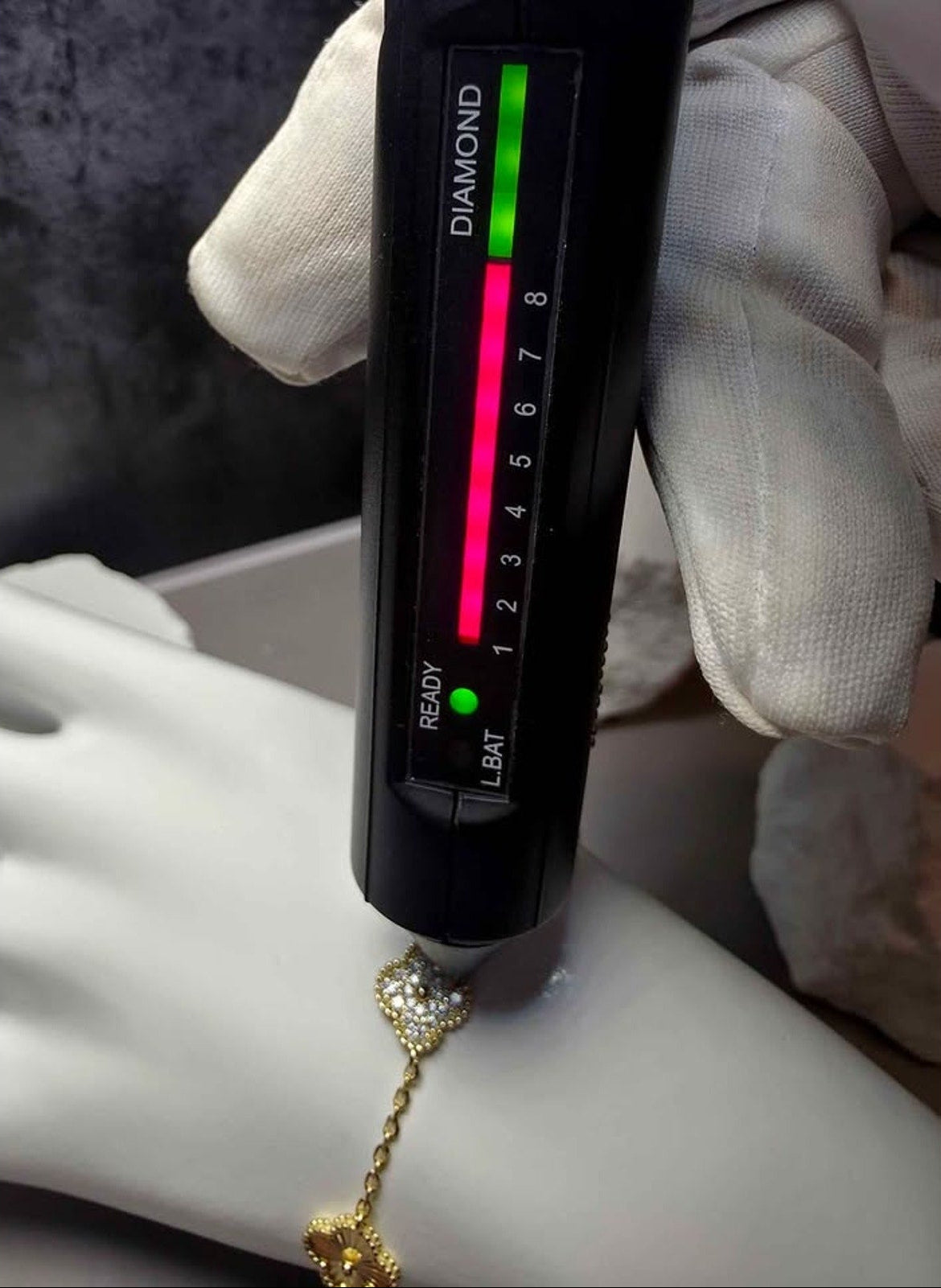 A close-up of a hand wearing a white glove holding a black diamond tester tool over a white mannequin hand, with a gold chain and clear gemstone pendant hanging below. The tester displays a colored diamond bar labeled DIAMOND and a scale from 1 to 8.