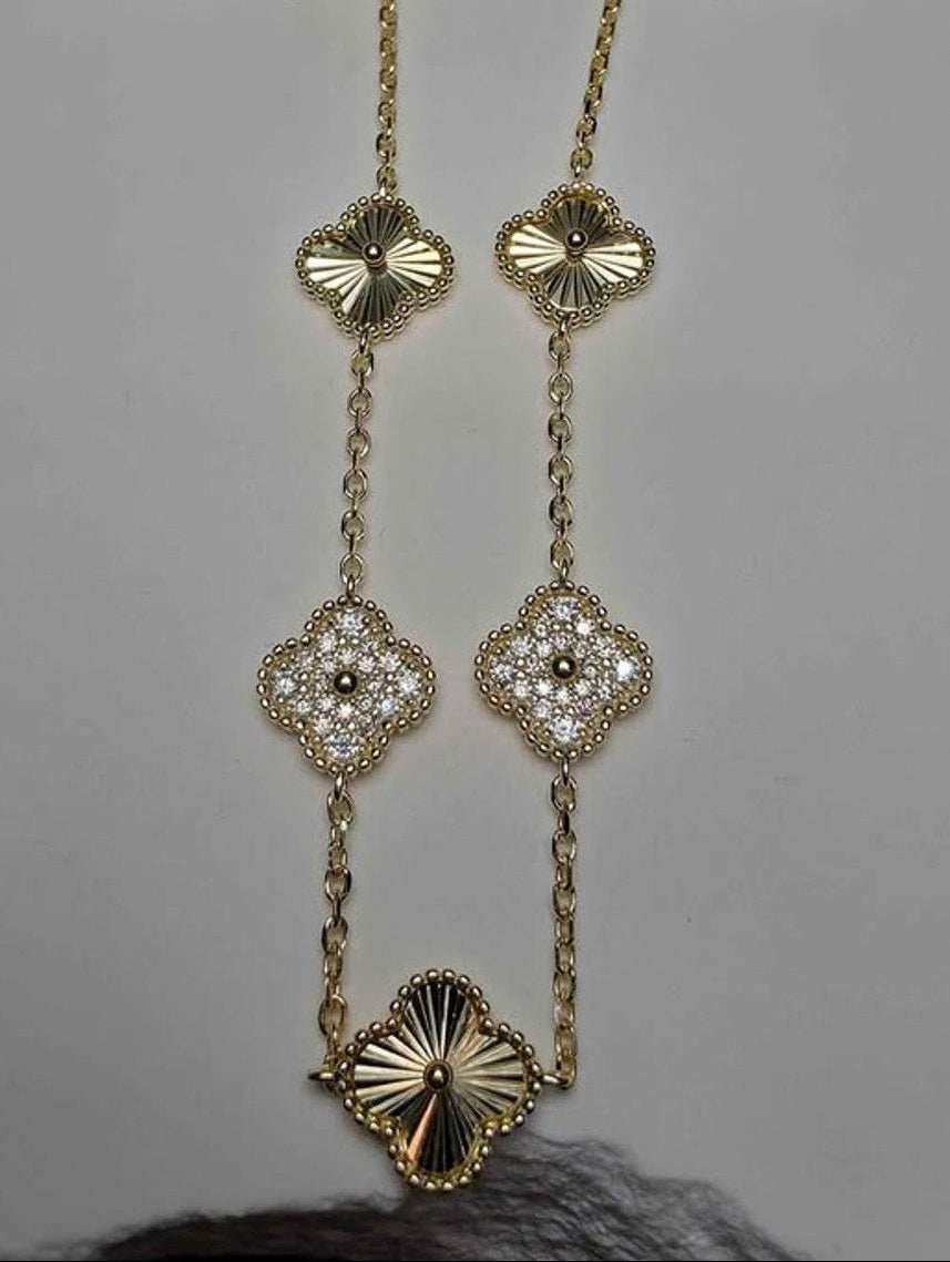 A gold-toned necklace with four clover-shaped pendants, each adorned with small clear stones, displayed on a light gray surface.