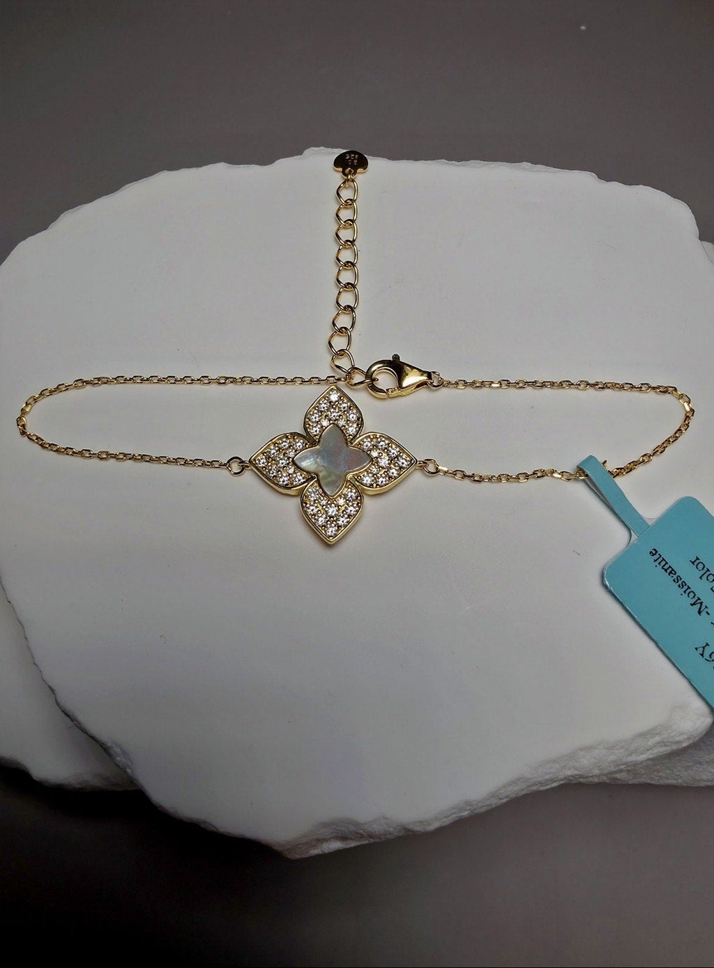 A gold chain bracelet with a four-leaf clover pendant set with clear stones and a central iridescent white inlay, displayed on a white stone slab with a light blue tag attached.