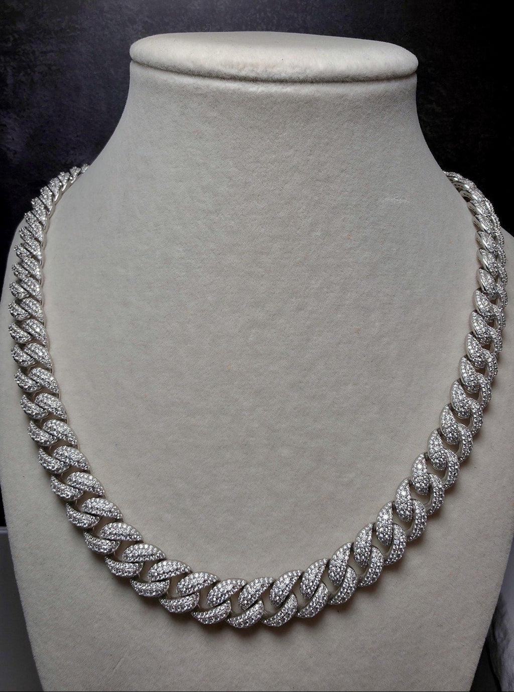 A thick silver Cuban link chain necklace, heavily embellished with small clear stones, displayed on a textured light gray velvet jewelry bust against a dark background.