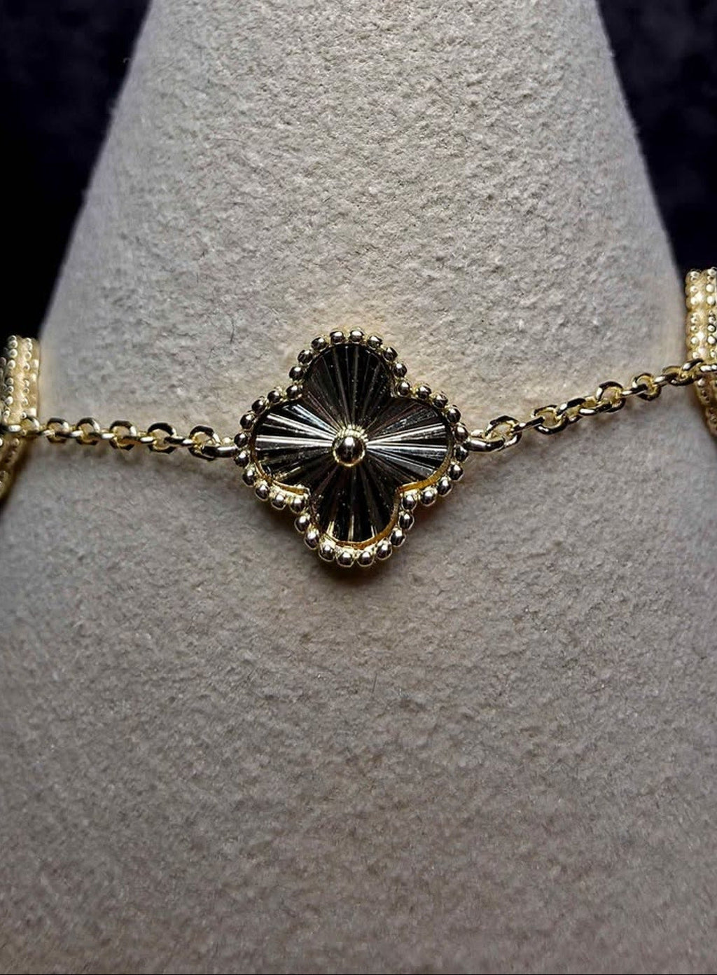 A gold-toned bracelet displayed on a textured light gray jewelry cone. The bracelet features a central clover-shaped motif with black and white striped detailing, surrounded by small clear stones, and connected by a gold chain.