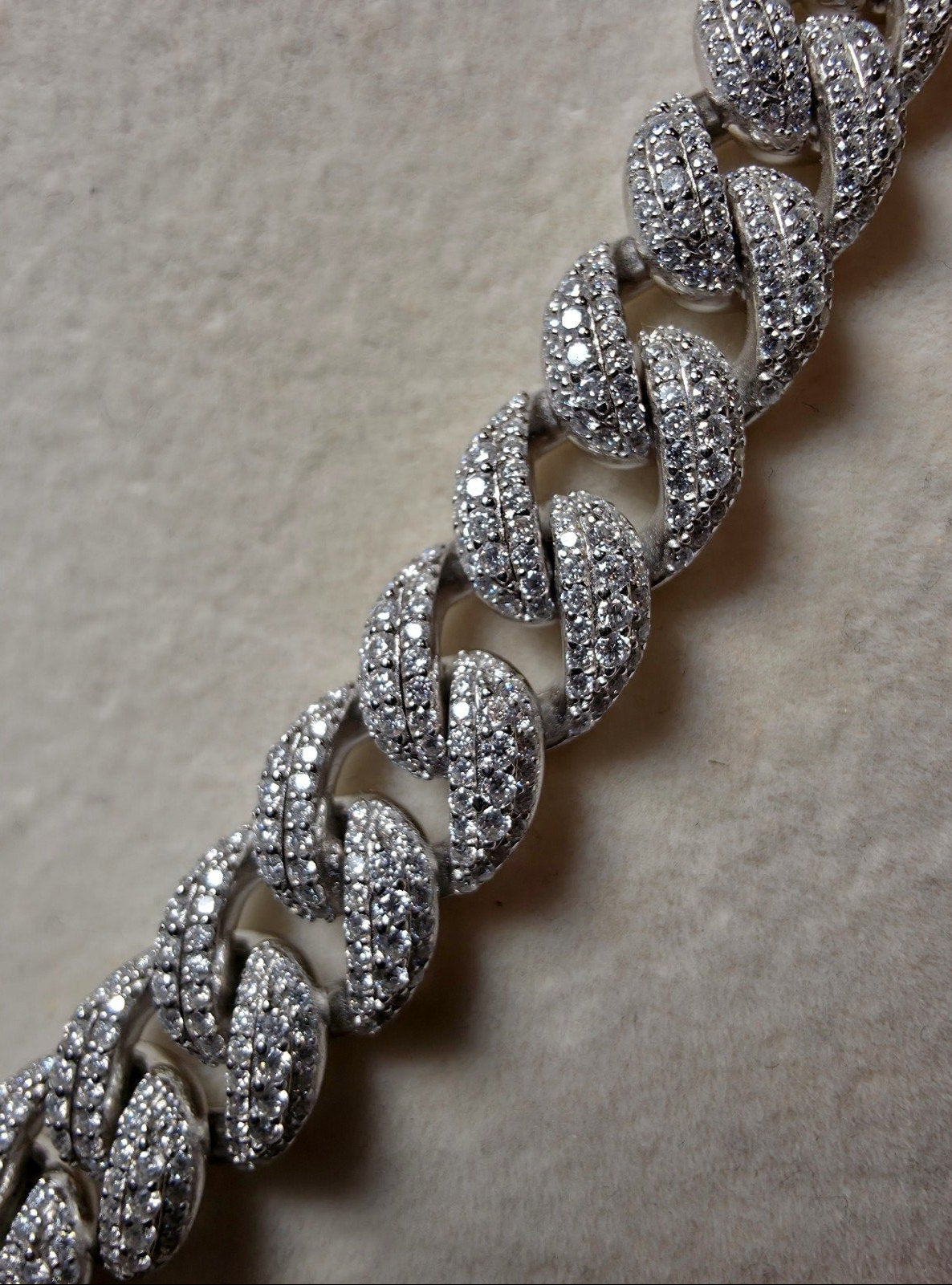 A close-up of a silver-toned Cuban link chain bracelet paved with clear cubic zirconia stones, resting on a textured beige surface.