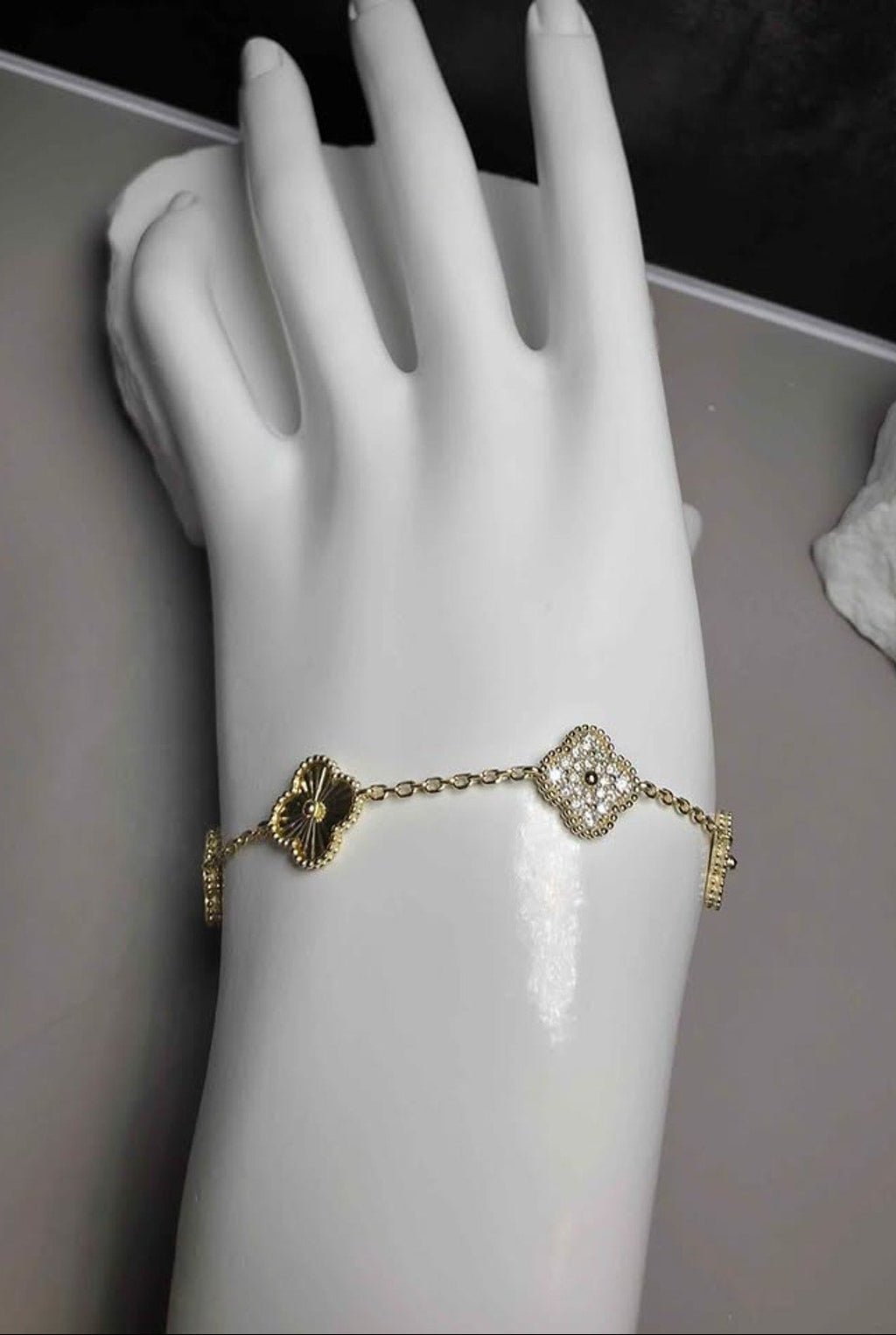 A gold-toned chain bracelet with two clover motifs, one dark gold and one clear crystal, displayed on a white mannequin hand.