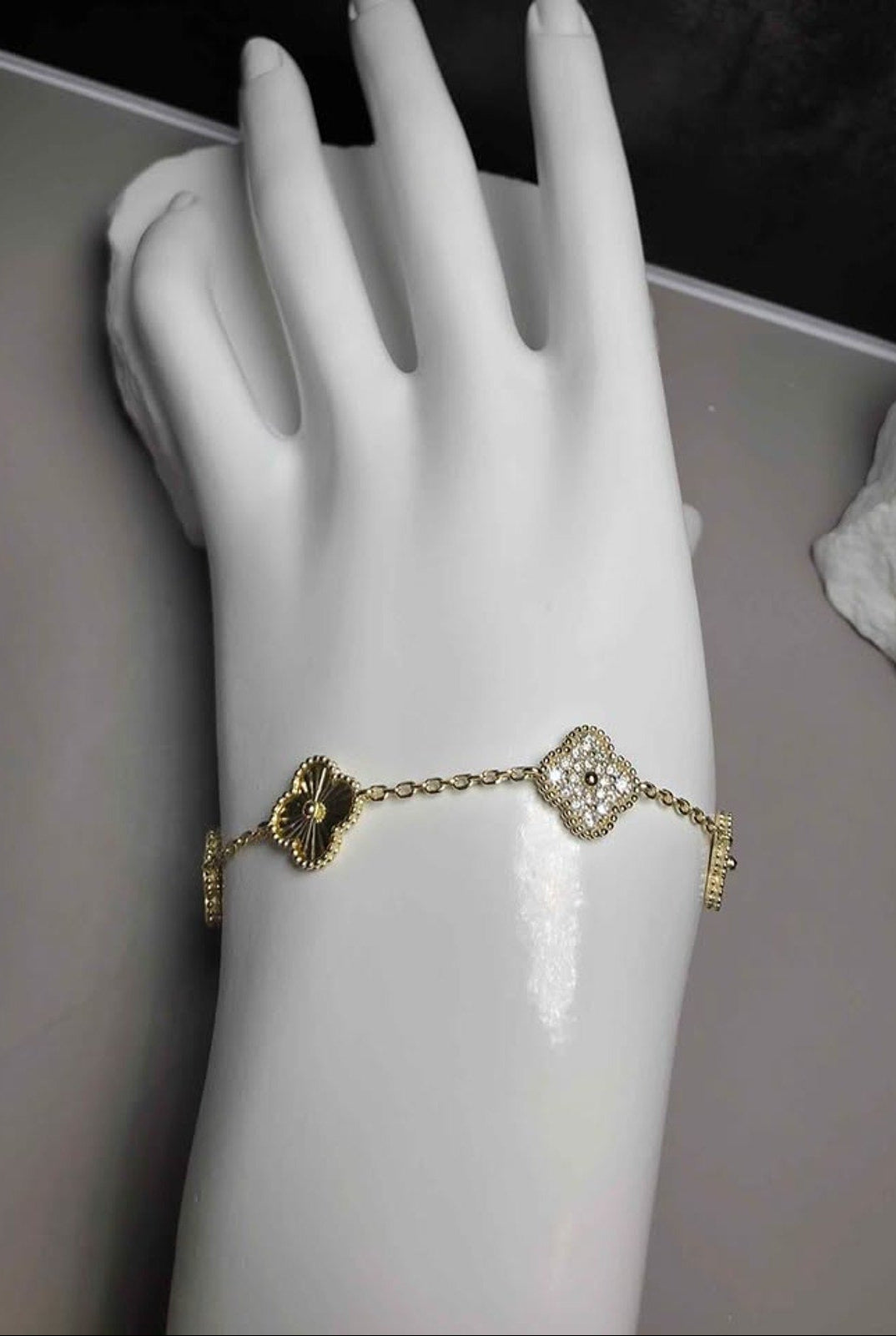 A gold-toned chain bracelet with two clover motifs, one dark gold and one clear crystal, displayed on a white mannequin hand.